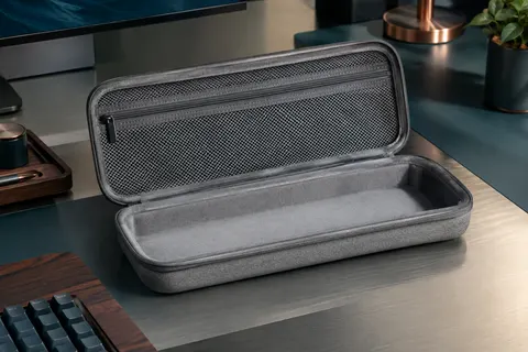 Étui rigide gris GEEKRIA ouvert, avec poche intérieure en filet zippée, posé sur un bureau sobre.